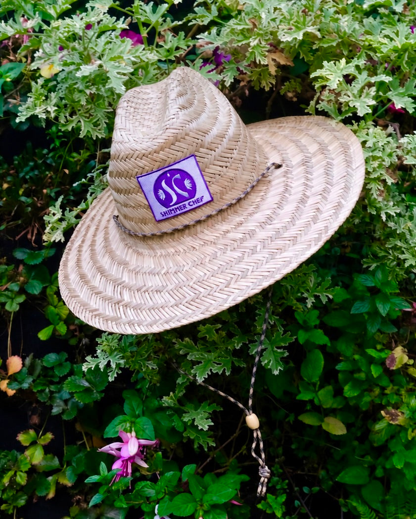 Shimmer Chef Lifeguard Hat, woven straw with lavender patch, resting on green foliage